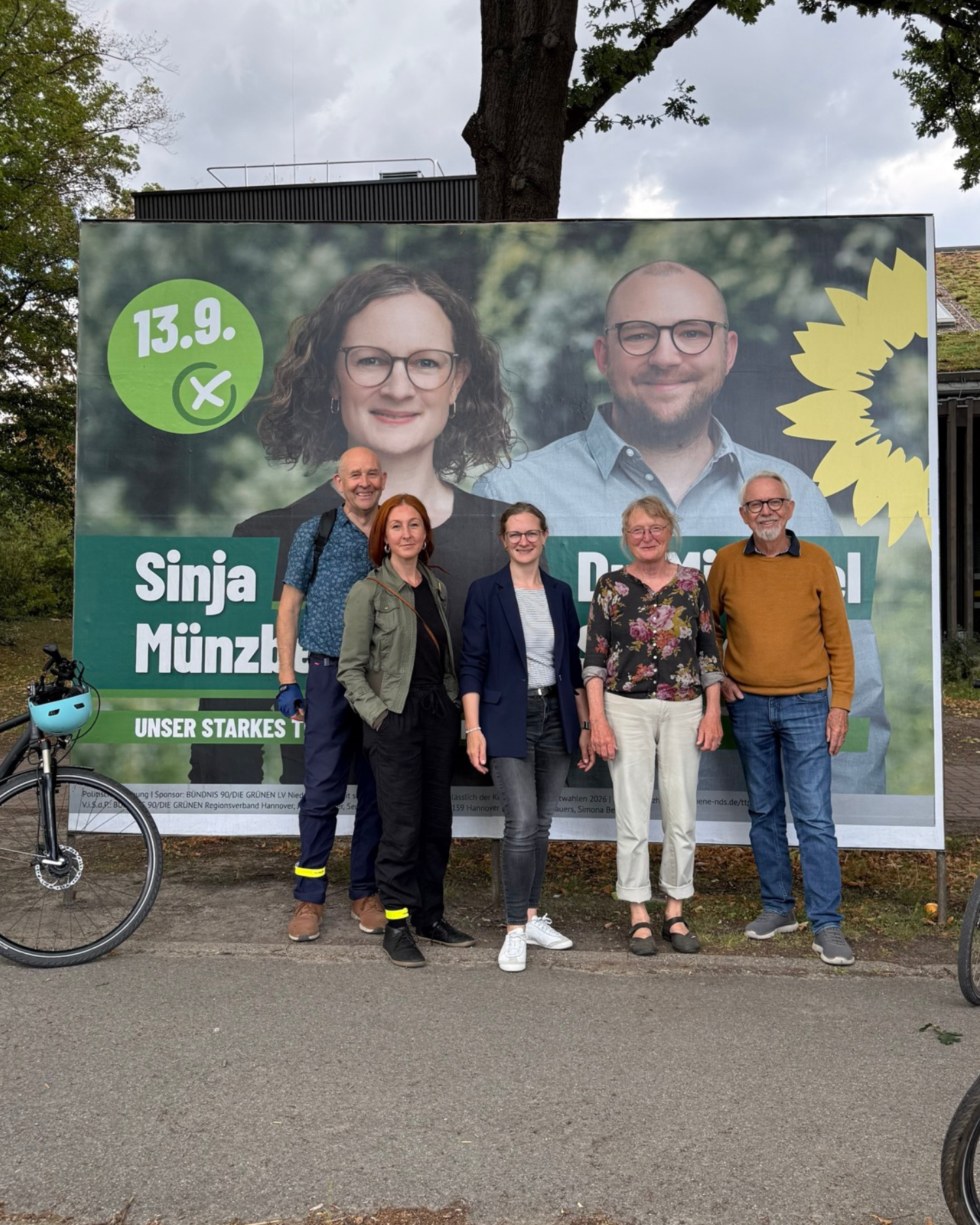 Fünf Personen stehen vor einem großen Wahlplakat mit Porträts einer Frau und eines Mannes sowie einem grünen Logo mit dem Datum '13.9.'