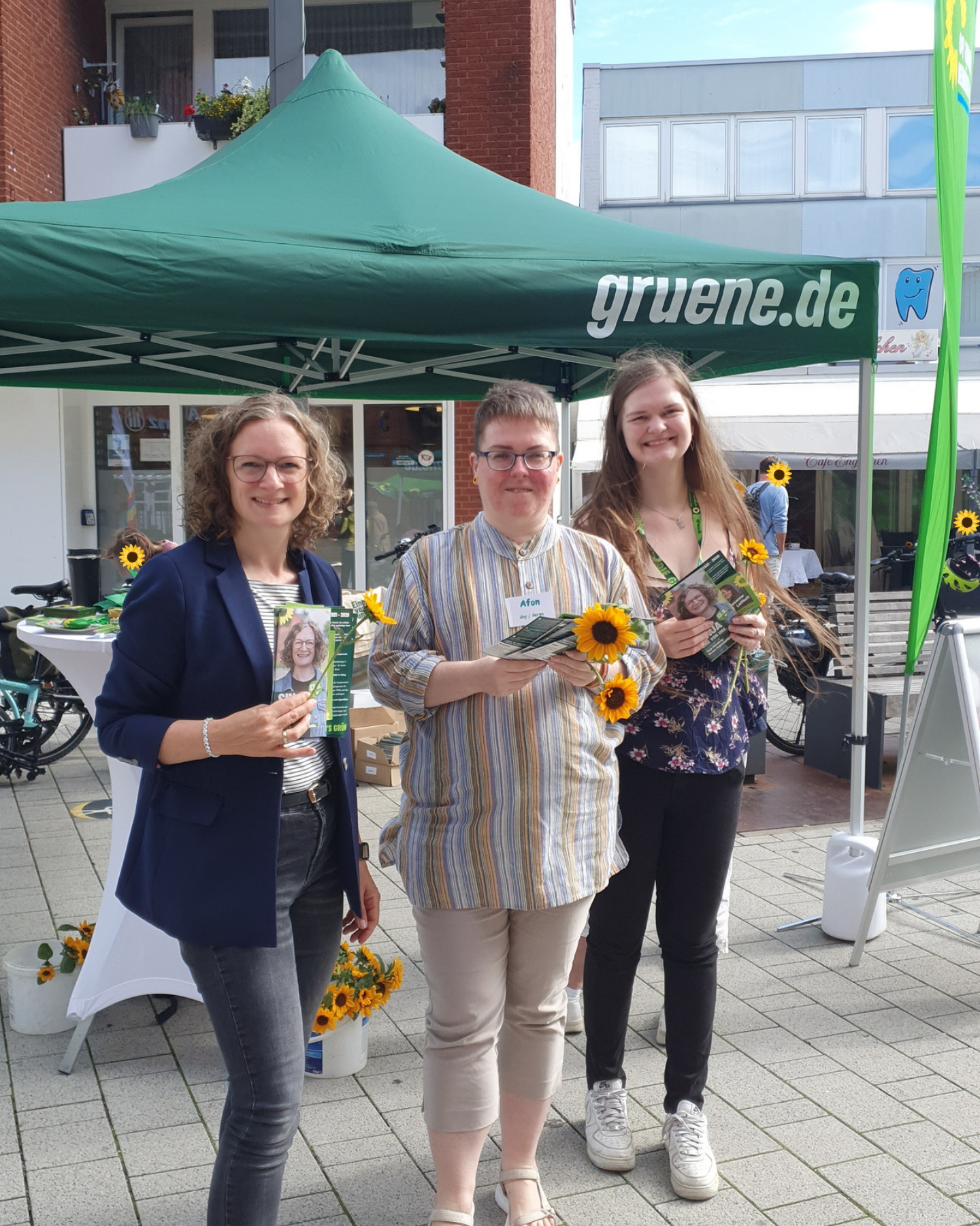 Drei lächelnde Frauen stehen vor einem grünen Pavillon mit der Aufschrift 'gruene.de' und halten Sonnenblumen und Flyer in der Hand